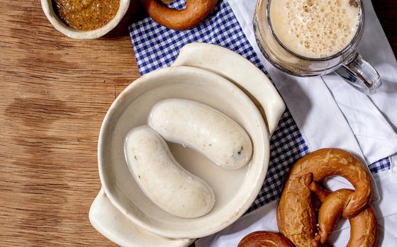 A round ceramic bowl with two white sausages in it, served with brown sweet mustard, a mug of dark beer and fresh pretzel bread on white and blue napkin over wooden background. 