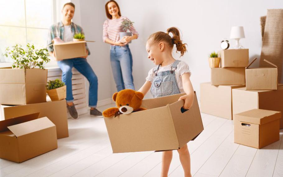 Little girl holding moving box with teddy bear popping out of the top and parents in background